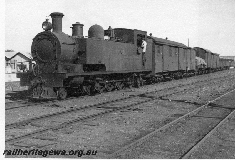 P02978
K class 35 steam locomotive on a goods train, driver on the footplate, front and side view, Kalgoorlie.
