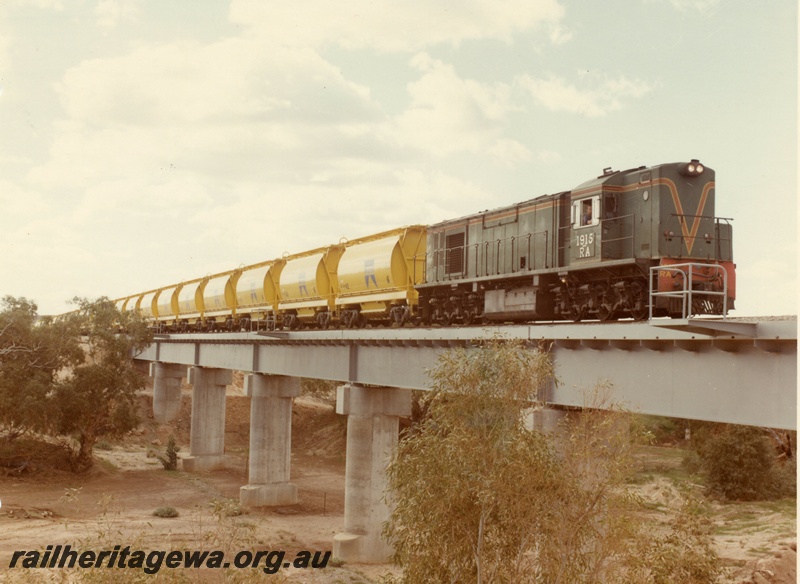 P03875
1 of 2, RA class 1915 diesel locomotive on mineral sands train, crossing the Irwin River bridge, running short hood first, side and front view, in green with red and yellow stripe livery, XE class bogie ilmenite hoppers.
