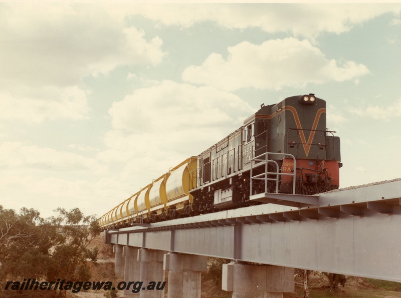 P03876
2 of 2, RA class 1915 diesel locomotive on mineral sands train, crossing the Irwin River bridge, running short hood first, side and front view, in green with red and yellow stripe livery, XE class bogie ilmenite hoppers.
