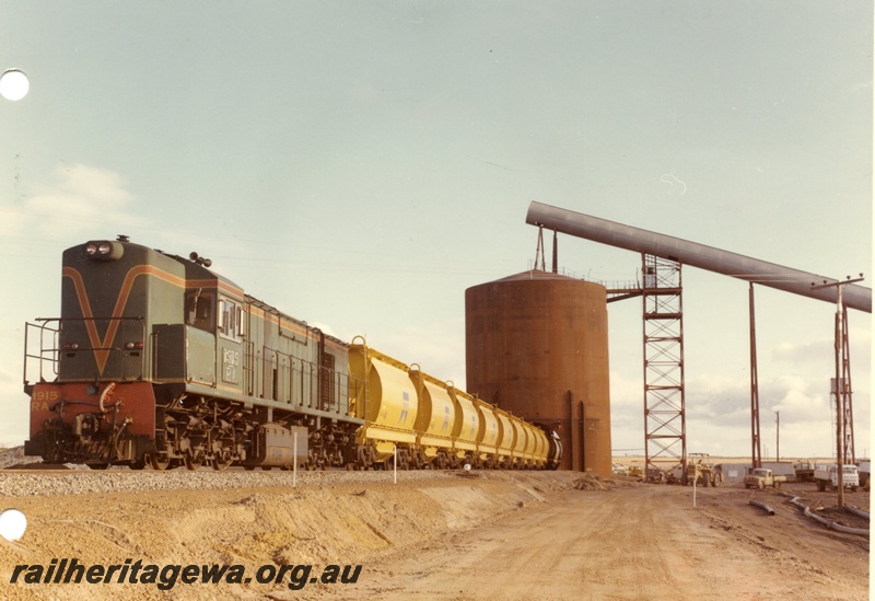 P03880
RA class 1915 diesel locomotive on mineral sands train, running short hood first, front and side view, in green with red and yellow stripe livery, loading XE class bogie ilmenite hoppers, Eneabba.
