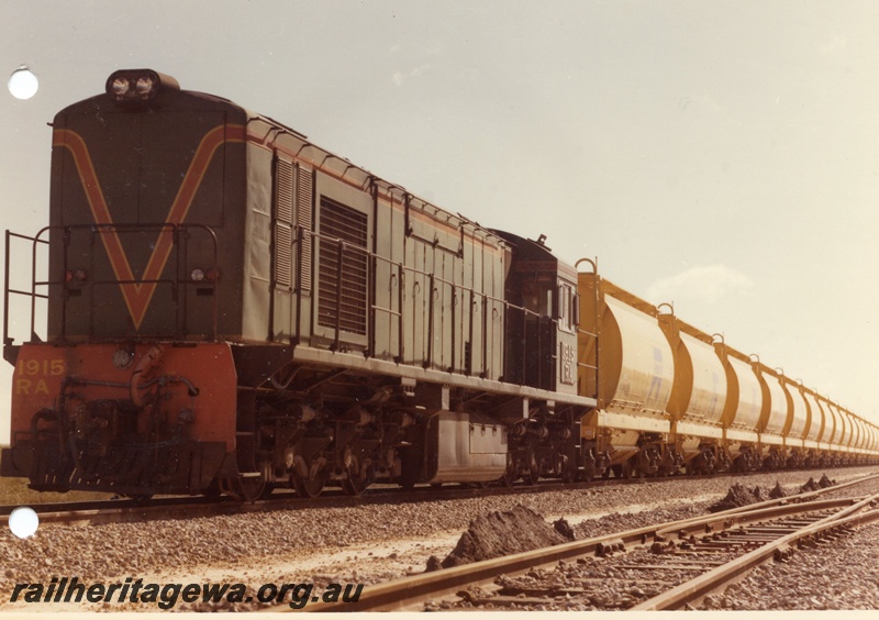 P03881
RA class 1915 diesel locomotive on mineral sands train, running long hood first, front and side view, in green with red and yellow stripe livery, XE class bogie ilmenite hoppers.

