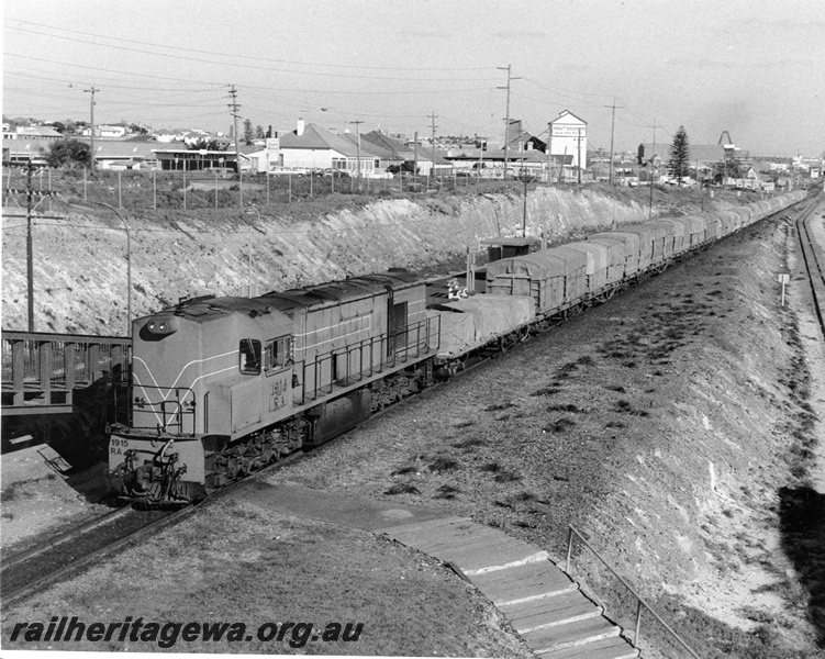 P10356
RA class 1915, Orange Blue and White Stripes on freight conveying ammonium nitrate and superphosphate loading passing through Leighton Station. Views of North Fremantle Container Terminal and Leighton Loco Depot in background. Narrow gauge track into Leighton Yard on right of picture, elevated photo taken from footbridge.
