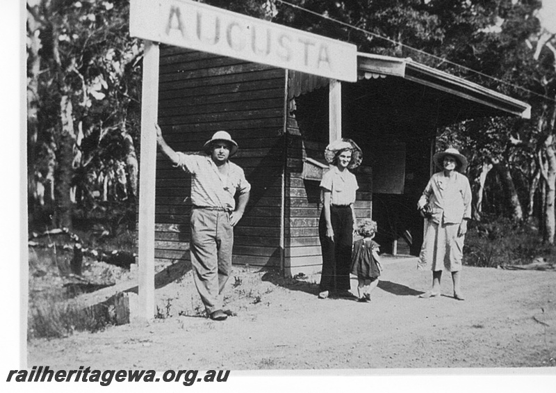 P15809
Station nameboard, shelter shed, Augusta, BB line, three adults and a small child in front of the nameboard
