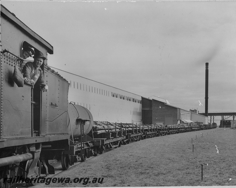 P15835
D type loco trailing a J class water tank wagon and a string of bolster wagons carrying steel products, BHP rolling mill at Kwinana, crew leaning out of the cab, view along the train

