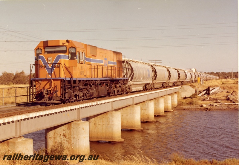P15895
N class 1877, in Westrail orange with blue and white stripe, on alumina train, crossing low concrete and steel bridge over river, front and side view
