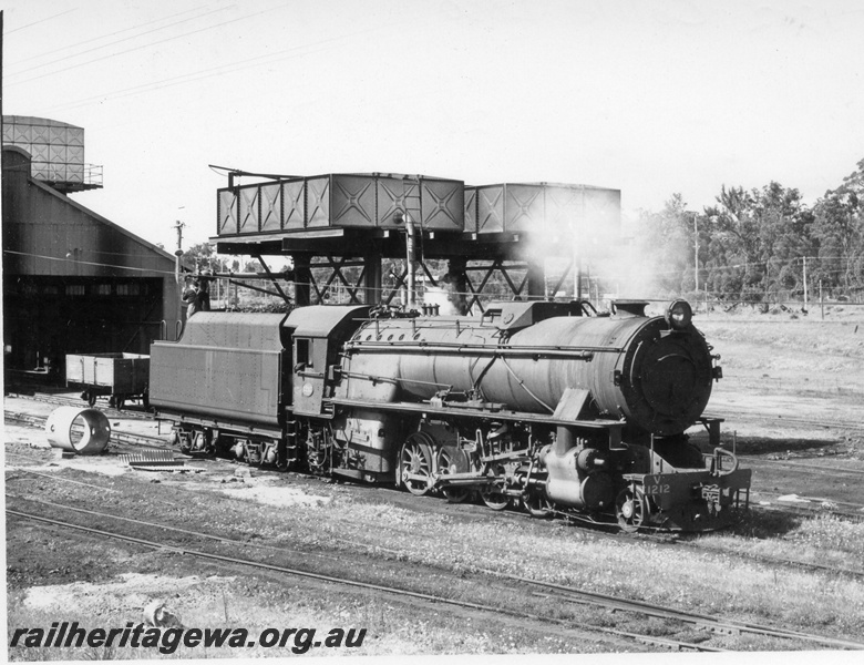 P17470
V class 1212 steam locomotive at Collie Loco with 2 water towers to right rear of locomotive. Loco sheds in background and an empty open goods wagon between shed and loco. BN line.
