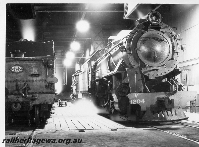 P17475
V class 1204 and PR 522 steam locomotives at Narrogin loco. GSR line. Front view of V class and rear view of tender of PR. Note the wooden planks in the shed flooring.
