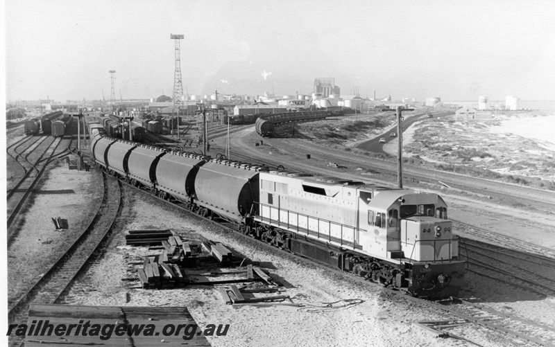 P18164
L class loco on loaded wheat train arriving Leighton, signals, rakes of goods carriages, beach, port of Fremantle in background, c1969
