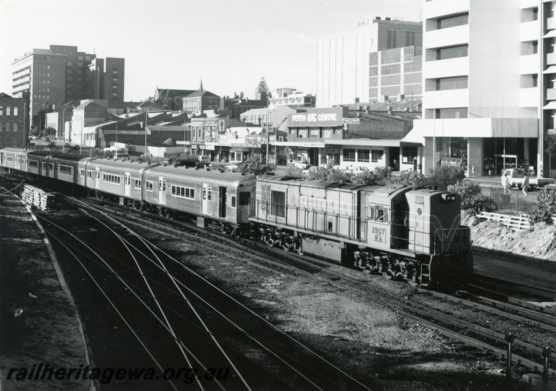 P18051
RA class 107, heading suburban passenger train comprising leased Queensland coaches, city buildings, arriving Perth City 
