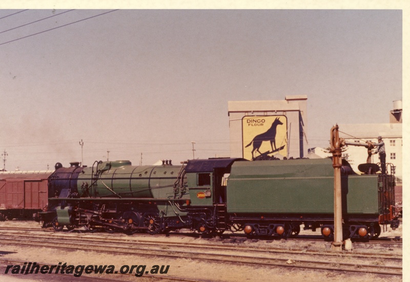 P18089
V class 1220, taking on water, water tower, Dingo Flour mill, sign on mill features black dingo on yellow background, Leighton, side and rear view, c1966
