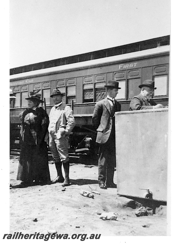 P19180
Four passengers including Sir John Forrest (extreme right) standing in front of the first Trans-Australian train from Port Augusta to Kalgoorlie during one of its stops, Commonwealth Railways (CR) first class sleeping car (part only), water tank, TAR line, track level view
