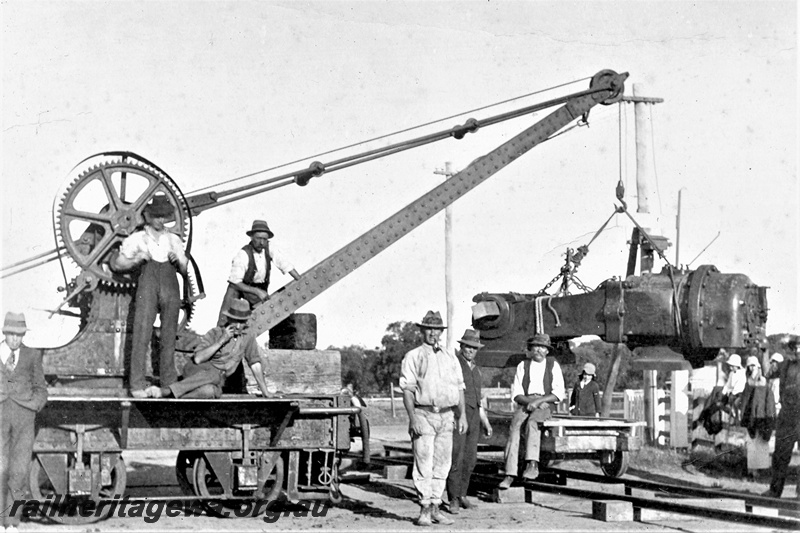P19697
Six wheel Cowans Sheldon rail mounted hand crane lifting an unidentified load, workers standing around, side view of part of the crane
