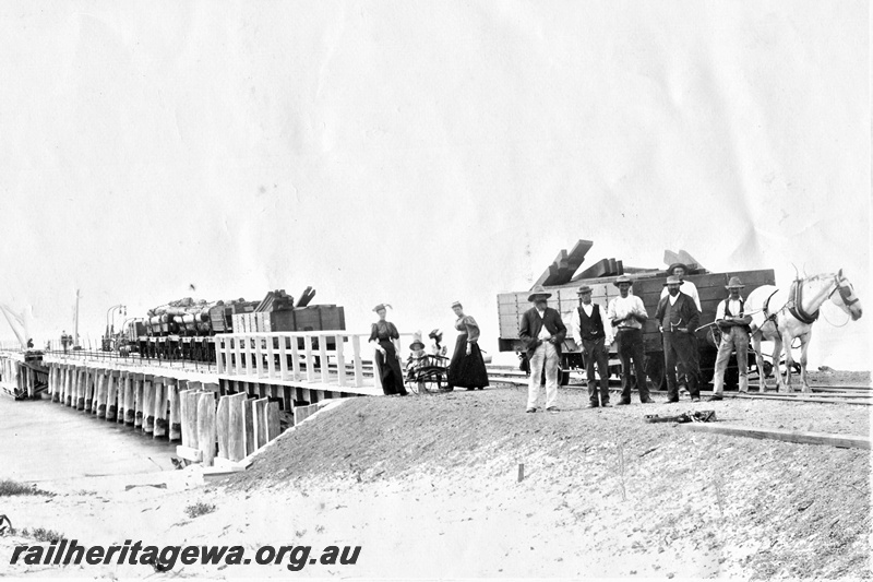 P19699
A horse drawing an R class bogie open wagon with a timber load, workers and two women and children in the picture, wagons on the jetty are another R class, I class bolster wagons and a G class wagon at the rear. Wagons are in the grey livery with black ironwork, loaded with sawn timber and logs. Photo taken of the Busselton jetty before the extension to the jetty in the 1890s
