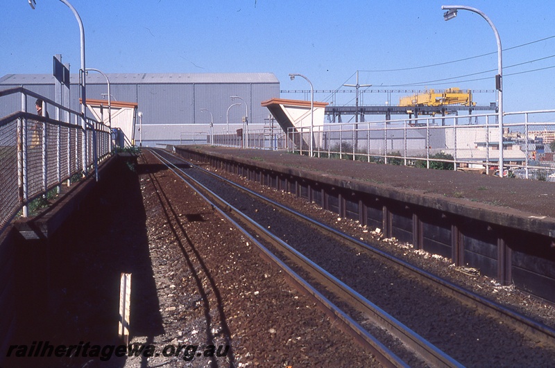 P19711
Platforms, shelters, dual gauge track, waiting passenger, shed and crane in background, North Fremantle, ER line, track level view
