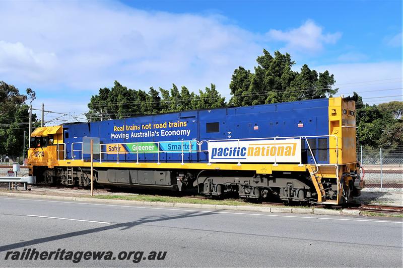 P21023
Pacific National NR class 14.in the blue and yellow livery with Real Trains Not Road Trains  moving Australia's Economy  Safer, Greener, Smarter  painted on the side of the loco, parked next to Railway Parade, Bassendean, side view. 
