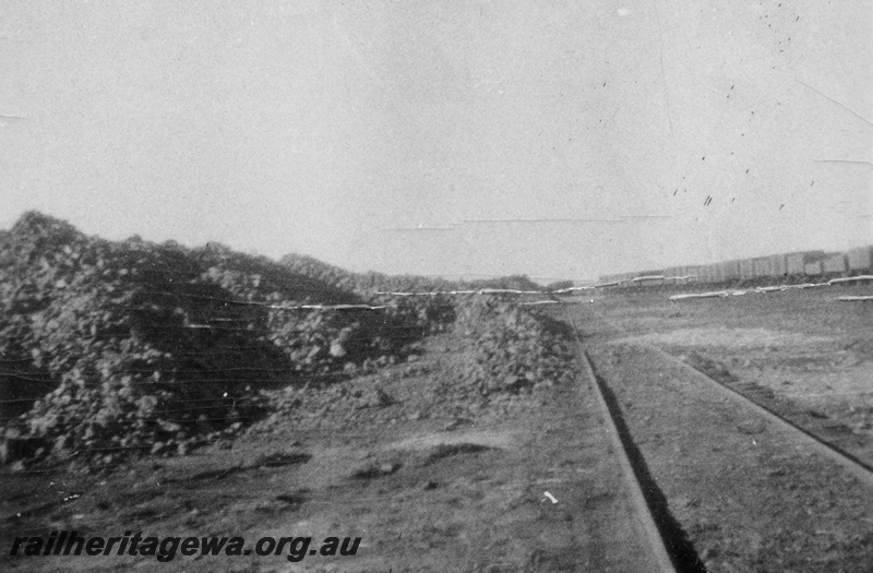 P21249
Siding, stockpiles of manganese ore next to the track, rake of open wagons on a track in the background, view along the siding, possibly Meekatharra, NR line
