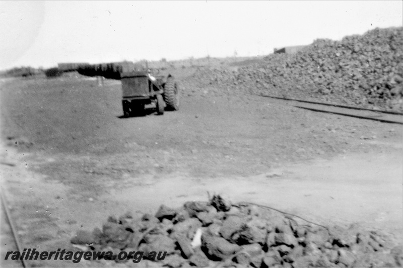 P21250
Siding, stockpiles of manganese ore next to the track, front end loader between the tracks, rake of open wagons on a track in the background, view along the siding, possibly Meekatharra, NR line
