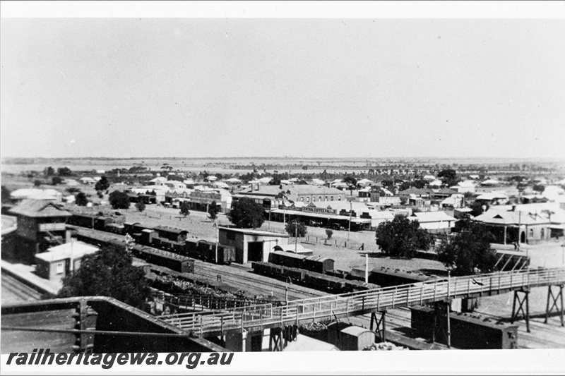 P21307
Station buildings, signal box, platform, pedestrian overpass, rakes of carriages, goods shed with soot mark over entry portal, town in background, Merredin, EGR line, view from elevated position
