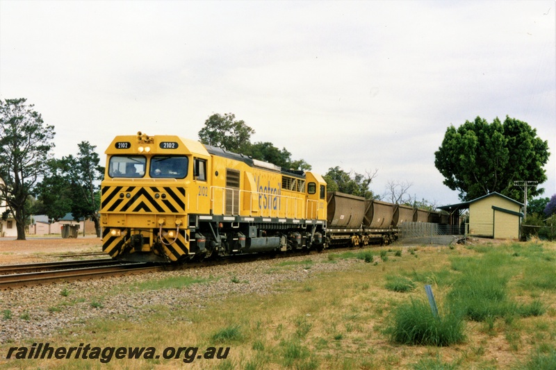 P21355
Westrail S class 2102 loco in the yellow with black chevrons livery heading an empty bauxite train past the Mundijong station building, SWR line, view along the train.
