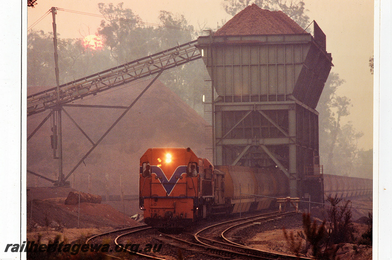 P22159
D class 1562, in Westrail orange with blue and white stripe livery, on woodchip train, loading woodchips, conveyor, overhead loader, Lambert, PP line, front and side view at track level
